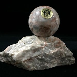 May include: A polished, spherical stone orb with pink, white, and grey marbling resting on a rough, unpolished base of the same stone. A circular gold sticker with black Japanese text is affixed to the top of the sphere, featuring the characters 特選.