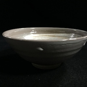 May include: A handmade ceramic bowl with a wide, shallow design. The bowl has a light brown exterior with subtle texture and a lighter interior. The rim is smooth, and the bowl sits on a small, rounded base. The background is black.
