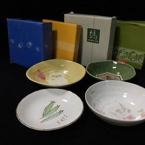 May include: Four ceramic bowls and four matching rectangular boxes. The bowls feature a yellow bowl with a white creature, a green bowl with a brown emblem, a white bowl with green plant illustrations, and a white bowl with pink floral patterns.