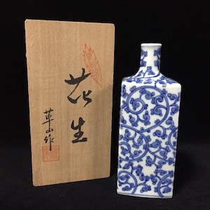 以下が含まれることがあります： 青と白の唐草模様が施された長方形の磁器製ボトル。ボトルの横には、黒い日本語の書道文字と赤い印が押された明るい茶色の木製収納箱が置かれています。