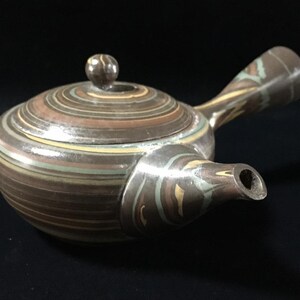 May include: A ceramic teapot with a side handle and a lid featuring a knob. The body displays a swirling, marbled pattern of brown, tan, cream, and muted green horizontal stripes that wrap around the entire vessel, including the spout and handle.