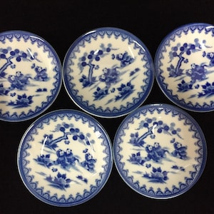 May include: A set of five round ceramic saucers featuring a white base with a blue painted pattern of stylized trees, foliage, and small figures. Each saucer has a decorative blue border with a repeating scalloped edge design around the rim.