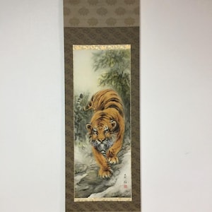 May include: A vertical Asian scroll painting featuring a detailed depiction of a tiger. The tiger, with orange and black stripes, is walking towards the viewer. The background includes trees and rocks. The scroll has a decorative top and bottom.