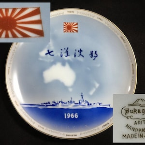 以下が含まれることがあります： 青と白のデザインのヴィンテージ日本の磁器プレート。プレートには、日の丸のエンブレム、船のシルエット、日本語の文字が描かれています。「Fukagawa Arita Handpainted Made in Japan」と1966年のマークが付いています。