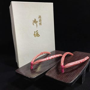 May include: A pair of dark brown wooden geta sandals with red fabric thongs featuring a white floral pattern. The sandals are positioned in front of a white rectangular box with gold Japanese calligraphy text printed on the front surface.