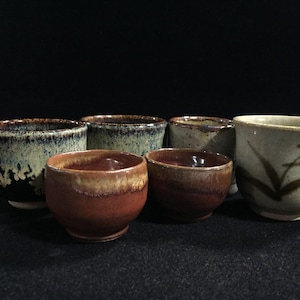Może przedstawiać: Zestaw sześciu ceramicznych czarek do herbaty o różnych wykończeniach szkliwa. Komplet zawiera naczynia o powierzchniach nakrapianych niebiesko-czarnych, jednolitych czerwonobrązowych i jasnobeżowych, z jedną czarką z malowanym brązowym wzorem liścia.