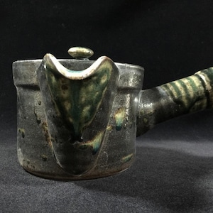 May include: A small, handcrafted ceramic teapot with a dark, textured finish. The spout and handle are accented with green and white glaze. The lid has a small, round knob. The teapot is designed for brewing tea.