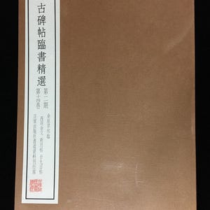 May include: A rectangular brown book cover featuring a white vertical label on the left side with black Chinese characters. The text includes the title 古碑帖臨書精選, volume information, and a small red square seal at the bottom left corner.
