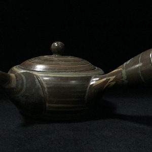 Può includere: Una teiera in ceramica con manico laterale, base marrone scuro e fasce orizzontali nei toni del beige, grigio e blu-verde. La teiera presenta un corpo arrotondato, un beccuccio corto, un coperchio abbinato con pomello e un manico largo e cavo.