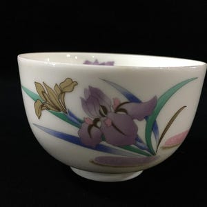 May include: A white ceramic bowl featuring a painted floral design of purple and tan irises with green and blue leaves. The bowl has a smooth, rounded shape with a small circular base, showcasing delicate botanical illustrations on its exterior surface.