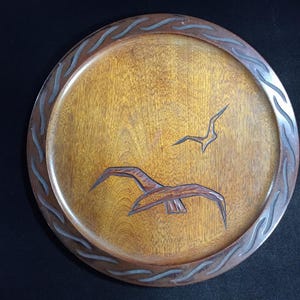 Op de afbeelding: Een ronde houten schaal met een gesneden ontwerp. De schaal heeft een lichtbruin midden met twee gesneden vogels in vlucht. De rand heeft een donkerbruine rand met een golfachtig patroon. Het is een decoratief item.
