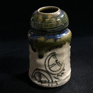 May include: A cylindrical ceramic vessel with a narrow neck and a rounded rim. The upper portion features a glossy green and blue glaze that drips down onto the off-white, textured lower body, which is decorated with black line drawings.