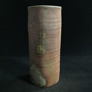 Könnte beinhalten: Eine zylindrische Keramikvase mit matter, erdbrauner Oberfläche. Der Körper weist feine horizontale Rillen sowie unregelmäßige, gesprenkelte Flecken in gedämpftem Grün und Anthrazit auf, was für eine texturierte und verwitterte Optik sorgt.
