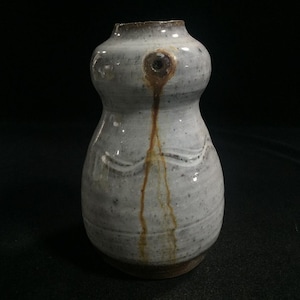 May include: A small, bulbous ceramic vase with a speckled light gray glaze. The vessel features a narrow neck, a rounded body, and a distinct brown drip pattern extending downward from a circular indentation near the top rim of the piece.