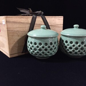 May include: Two light green celadon ceramic bowls with lids, featuring a perforated lattice pattern on the sides. The bowls are positioned in front of a rectangular wooden storage box tied with a dark brown ribbon, which has Japanese text.