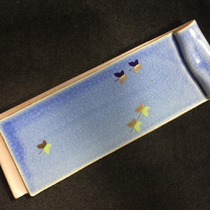 以下が含まれることがあります： ひび割れのある釉薬と巻き上げられた縁を持つ、長方形の薄い青色のセラミックトレイ。 金、紫、黄色の小さな、様式化された蝶のデザインで装飾されています。 トレイには薄いタン色のボーダーがあります。