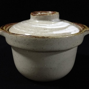 Op de afbeelding: Een kleine, ronde, gespikkelde beige keramische pot met bijpassend deksel. De pot heeft twee kleine handvatten aan de zijkanten en een bruine rand. Het deksel heeft een centrale knop en een wit, wervelend glazuurpatroon op de bovenkant.