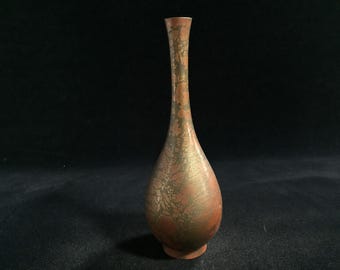 Puede incluir: Un jarrón alto y esbelto con cuello estrecho y base bulbosa. La superficie presenta un acabado moteado y texturizado en tonos cobre apagado, marrón rojizo y bronce oscuro, creando una apariencia orgánica y desgastada en todo el exterior.