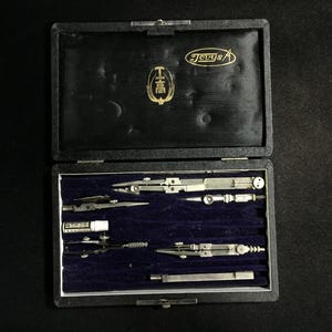 Op de afbeelding: Een set vintage tekeninstrumenten in een zwarte rechthoekige doos met een donkerblauwe fluwelen binnenkant. De set bevat diverse metalen passers, een kleine houder voor potloodvullingen en een metalen verlengstuk. Het deksel bevat de tekst Jevys.