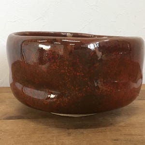 May include: A round, hand-formed ceramic bowl with a glossy, deep reddish-brown glaze. The surface features subtle variations in color and texture, with a smooth, reflective finish and a slightly irregular, organic rim shape.