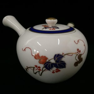 May include: A white ceramic teapot with a side handle and a lid featuring a blue rim. The body is decorated with a painted botanical pattern of red berries and blue, orange, and brown leaves on thin, winding stems. The lid has a gold-colored knob.