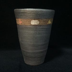 Op de afbeelding: Een donkergrijze, getextureerde keramische beker met een taps toelopende vorm. Een horizontale band van bruine klei omcirkelt het bovenste gedeelte, voorzien van kleine, handgeschilderde accenten in rood, zilver en goud, waaronder een driehoek en een rechthoek.