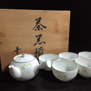 May include: A white ceramic teapot with a side handle and lid, decorated with green bamboo-like motifs and a blue rim, accompanied by four matching white tea cups and a wooden storage box featuring black Japanese calligraphy characters.