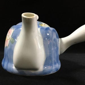 以下が含まれることがあります： 白い注ぎ口と取っ手が付いた陶器のティーポット。本体は水色で、ピンクと黄色の花の模様が描かれています。ティーポットは丸みを帯びた形で、黒い面に置かれています。
