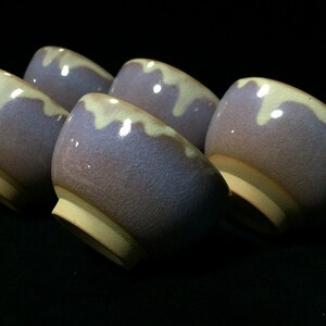 Peut inclure: Un ensemble de cinq petits bols en céramique présentant une glaçure violet lavande avec un motif de coulures crème le long du bord. Chaque bol possède une texture lisse et craquelée ainsi qu'une base beige non émaillée, disposés ensemble.