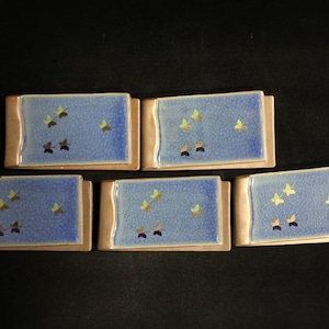 Könnte beinhalten: Ein Set aus fünf rechteckigen Essstäbchenablagen aus Keramik mit einer texturierten, blauen Glasuroberfläche in Craquelé-Optik. Jedes Stück ist mit kleinen, goldfarbenen Schmetterlingsmotiven verziert und hat einen hellbraunen, unglasierten Rand.