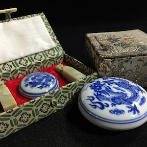May include: Two round white porcelain containers with blue dragon designs, accompanied by two rectangular pale green stone seals. One container rests inside a fabric-covered box with a red velvet interior, while the other sits beside a matching box.