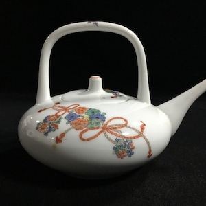 May include: A white ceramic teapot featuring a high arched handle, a tapered spout, and a lid. The body is decorated with a delicate pattern of orange, blue, and green floral clusters connected by thin, intertwined orange rope-like motifs.