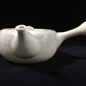 Może przedstawiać: Biały ceramiczny czajnik z długim, zakrzywionym uchwytem i dzióbkiem. Czajnik ma zaokrąglony korpus i mały uchwyt na górze. Czajnik jest przeznaczony do nalewania herbaty.