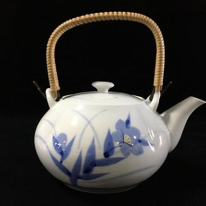 以下が含まれることがあります： 手描きの青い花柄と緑の葉が描かれた白い陶磁器製の急須。丸みを帯びた本体に、共蓋、短い注ぎ口、そして持ち手には薄茶色の籐のような素材が巻かれたデザインで、全体的に滑らかな質感の陶器製ティーポットです。