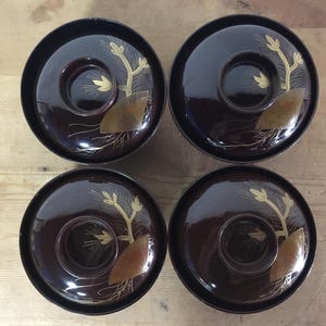 Könnte beinhalten: Ein Set aus vier dunkelbraunen lackierten Schalen mit passenden Deckeln. Jeder Deckel ist mit einem goldenen Motiv aus einem Fächer und Kiefernnadeln verziert. Die Schalen haben eine glatte, glänzende Oberfläche und einen runden Griff.