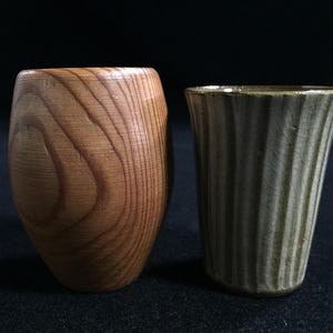 Könnte beinhalten: Zwei handgefertigte Trinkgefäße. Eines ist ein Holzbecher mit natürlicher Holzmaserung. Das andere ist ein Keramikbecher mit gerippter Textur und hellgrüner Glasur. Beide Becher werden vor einem dunklen Hintergrund präsentiert.