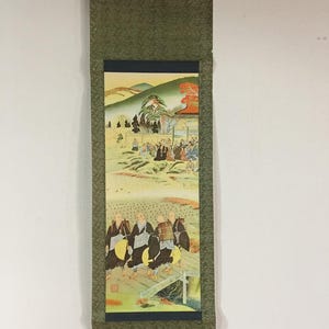 May include: A vertical Japanese hanging scroll featuring a detailed painting of a group of monks walking across a wooden bridge in the foreground, with a landscape of rolling hills, trees, and a gathering of people in the background, framed in green.