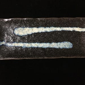 May include: A rectangular ceramic serving plate with a dark, speckled black and gray base. The surface features two horizontal, textured white and light blue brushstroke patterns that span across the center of the dish.