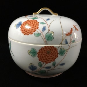 May include: A white porcelain lidded bowl featuring a hand-painted floral pattern with orange chrysanthemum-like flowers, green leaves, and blue accents. The lid has a gold-trimmed handle, and the overall design includes delicate gold line work.