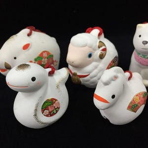 Puede incluir: Una colección de cinco figuras de animales de cerámica blanca que incluye un jabalí, una oveja, un perro, una serpiente y un caballo. Cada figura presenta detalles pintados, patrones florales coloridos y un cordón rojo en la cabeza.