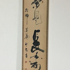 May include: A vertical hanging scroll featuring black ink calligraphy on a light tan paper background. The scroll is mounted on a textured brown fabric border with wooden rollers at the top and bottom. The artwork displays bold, expressive brushstrokes.