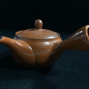 May include: A brown ceramic teapot with a unique handle design. The teapot has a lid with a small knob and a spout. The handle is a large, open loop. The teapot is likely used for brewing tea.