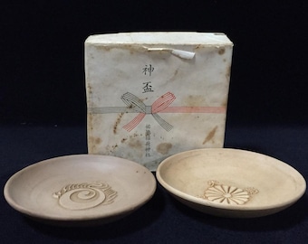 May include: Two round ceramic saucers with embossed designs, one featuring a spiral pattern and the other a chrysanthemum crest, placed in front of a square white box with Japanese text and a printed red and green ribbon graphic.