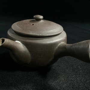 May include: A brown ceramic teapot with a lid and a handle. The teapot has a spout and a small knob on the lid. The teapot is likely used for brewing tea.