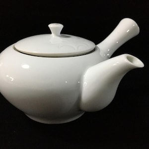 May include: A white ceramic teapot with a side handle and a matching lid. The teapot features a smooth, glossy finish and a rounded body shape with a short, curved spout. The design is simple and functional, showcasing a clean, minimalist aesthetic.