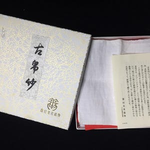 May include: An open box with a white and gold patterned lid, revealing a white cloth and a pamphlet. The box has Japanese text on the lid. The interior of the box is lined with red fabric.