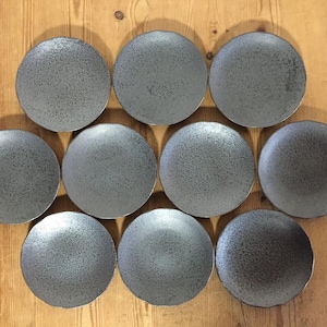 May include: A set of ten circular plates featuring a dark gray, speckled, textured finish. The plates are arranged in a grid-like pattern, each displaying a consistent matte appearance with subtle variations in the density of the dark speckles.