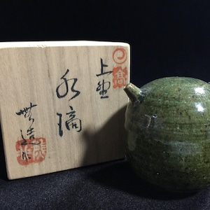 Könnte beinhalten: Ein runder, grün glasierter Keramik-Wassertropfer mit einem kleinen Ausguss, platziert neben einer quadratischen Aufbewahrungsbox aus Holz. Die Box zeigt schwarze japanische Kalligrafiezeichen und zwei rote Stempel auf der Vorderseite.