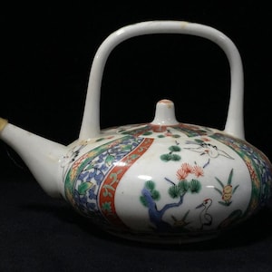May include: A white ceramic teapot featuring a high arched handle and a long spout. The body is decorated with colourful hand-painted motifs including green pine trees, flying cranes, and floral patterns arranged in segmented panels around the exterior.