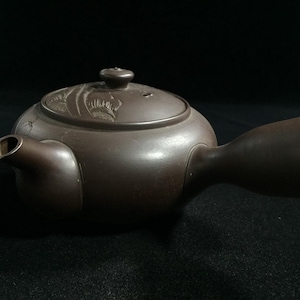 Könnte beinhalten: Eine braune Keramik-Teekanne mit seitlichem Griff und Deckel. Der Deckel hat ein Blumendesign. Die Teekanne hat einen Ausguss und einen breiten Griff. Der Hintergrund ist schwarz.